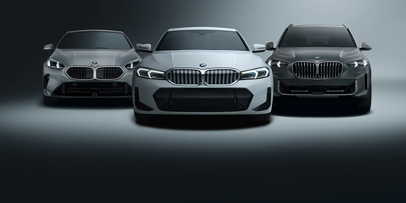 Front view of three BMW cars in a row, center white, in a dark studio setting with headlights on.